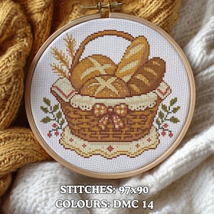 May include: A cross-stitch embroidery hoop featuring a basket filled with various types of bread. The basket is brown with a decorative bow, and the bread is in shades of tan and brown. The text at the bottom reads "STITCHES: 97x90 COLOURS: DMC 14."