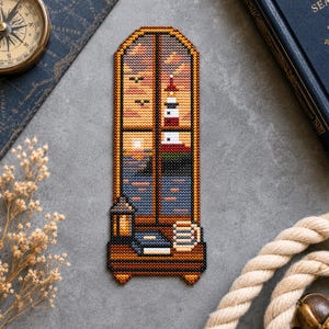 May include: A rectangular cross-stitch bookmark featuring a window view of a lighthouse at sunset over the ocean. The scene includes a lantern, a book, and a mug on a wooden ledge. The design uses shades of orange, blue, brown, and white thread.