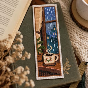 Può includere: Un segnalibro rettangolare a punto croce raffigurante una tazza di caffè fumante su un davanzale. Il disegno mostra una vista esterna piovosa attraverso una finestra con toni blu, una pianta verde e una tazza bianca con un piccolo motivo floreale.