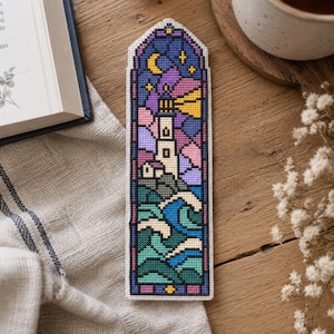 May include: A rectangular cross-stitch bookmark featuring a stained-glass style design of a lighthouse on a rocky shore at night. The scene includes a yellow crescent moon, stars, a beam of light, and stylised blue and green ocean waves.