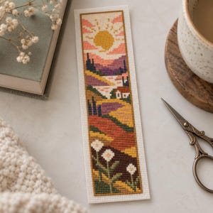 Può includere: Un segnalibro rettangolare a punto croce raffigurante un paesaggio con un sole giallo, cielo rosa, colline viola, una piccola casa e un sentiero tortuoso. Il primo piano mostra campi verdi e fiori bianchi, bordato da un bordo in tessuto bianco.