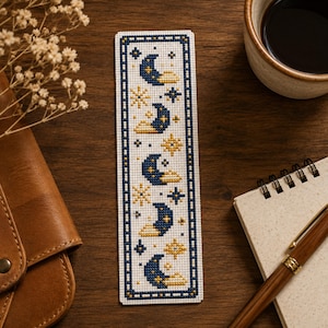 May include: A rectangular cross-stitch bookmark featuring a repeating pattern of dark blue crescent moons and yellow stars on a white fabric background, enclosed within a dark blue stitched border that frames the celestial design vertically.