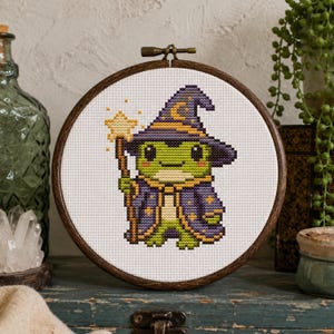 May include: A cross-stitch embroidery in a circular wooden hoop featuring a green frog wearing a purple wizard hat and robe with yellow star accents. The frog holds a yellow star-topped wand against a white fabric background.
