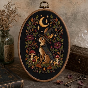 May include: An oval wooden embroidery hoop containing a cross-stitch design on black fabric. The image features a brown rabbit wearing a dark cloak and pointed hat, surrounded by colourful mushrooms, autumn leaves, flowers, and a crescent moon.