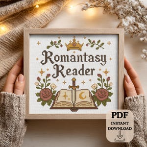 May include: A framed cross-stitch design with the words "Romantasy Reader" in brown thread. The design includes a crown, sword, open book, and floral elements in shades of brown, green, and pink. The frame is light brown.