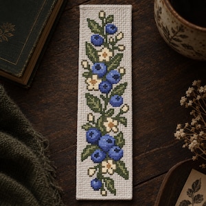 Op de afbeelding: Een rechthoekige boekenlegger met een kruissteekpatroon van blauwe bosbessen, groene bladeren en kleine witte bloemetjes op een lichtbeige stoffen achtergrond. Het borduurwerk is verticaal gecentreerd en toont een botanisch motief.
