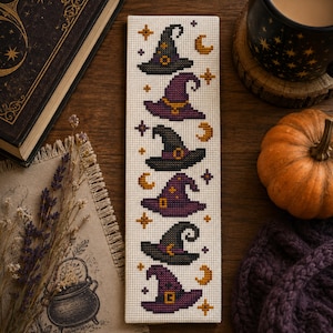 May include: A rectangular cross-stitch bookmark on white Aida fabric featuring a vertical pattern of six alternating purple and black witch hats, accented with small orange crescent moons and four-pointed stars in orange and dark purple.
