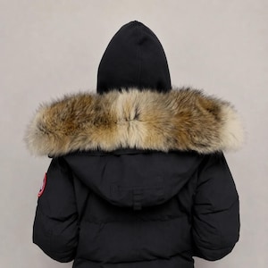 Puede incluir: Una parka de invierno negra con capucha y un grueso borde de piel en tonos marrones y tostados. La chaqueta presenta un diseño acolchado y un parche circular bordado en rojo, blanco y azul en la manga superior, con un logotipo detallado.