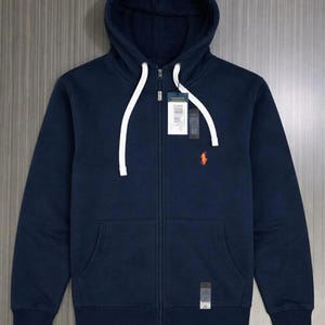 Pu&ograve; includere: Felpa con cappuccio blu navy con zip e tasca a marsupio. La felpa ha un cordino bianco e un piccolo logo arancione sul petto. Un'etichetta &egrave; attaccata vicino alla cerniera.