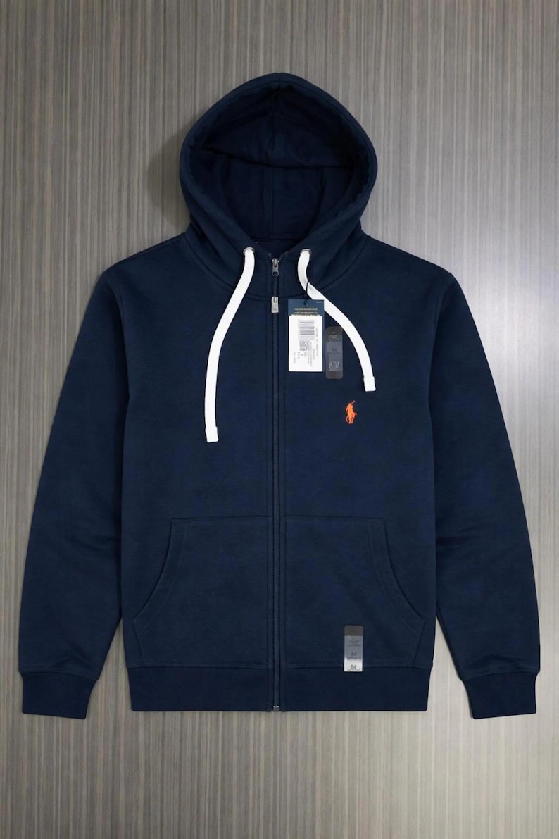 Pu&ograve; includere: Felpa con cappuccio blu navy con zip e tasca a marsupio. La felpa ha un cordino bianco e un piccolo logo arancione sul petto. Un'etichetta &egrave; attaccata vicino alla cerniera.