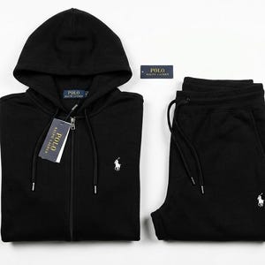 May include: Black tracksuit set comprising a hooded zip-up top and matching joggers. Both items feature a white logo. The top has a tag attached. The set is presented on a white backdrop.