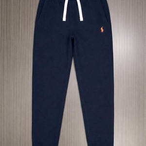 Pu&ograve; includere: Pantaloni della tuta blu navy con coulisse bianca e logo arancione sulla gamba sinistra. I pantaloni hanno due tasche laterali e polsini a coste alle caviglie. Il materiale sembra essere un tessuto morbido e confortevole.