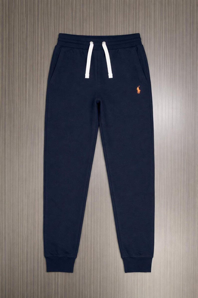 Pu&ograve; includere: Pantaloni della tuta blu navy con coulisse bianca e logo arancione sulla gamba sinistra. I pantaloni hanno due tasche laterali e polsini a coste alle caviglie. Il materiale sembra essere un tessuto morbido e confortevole.
