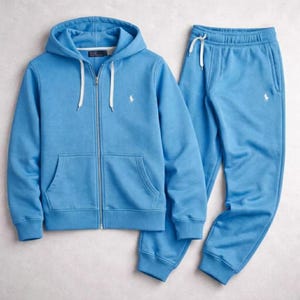 May include: A light blue matching sweatsuit set. The set includes a zip-up hoodie with a kangaroo pocket and drawstring, and matching sweatpants with a drawstring. Both pieces feature a small white logo on the chest. The hoodie has a white drawstring.