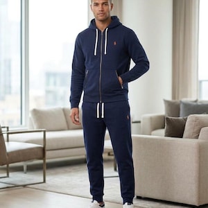 Pu&ograve; includere: Una tuta blu navy con una felpa con cappuccio con zip e pantaloni della tuta abbinati. La felpa con cappuccio ha un cordino bianco e un piccolo logo arancione sul petto. I pantaloni della tuta hanno la vita con coulisse e un logo abbinato. La persona indossa scarpe da ginnastica bianche.