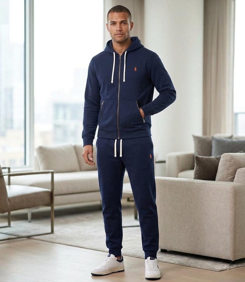 Pu&ograve; includere: Una tuta blu navy con una felpa con cappuccio con zip e pantaloni della tuta abbinati. La felpa con cappuccio ha un cordino bianco e un piccolo logo arancione sul petto. I pantaloni della tuta hanno la vita con coulisse e un logo abbinato. La persona indossa scarpe da ginnastica bianche.
