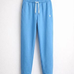 May include: Light blue sweatpants with a drawstring waist and a small white logo on the left leg. The pants have side pockets and are made of a soft material. The sweatpants are hanging on a wooden hanger.