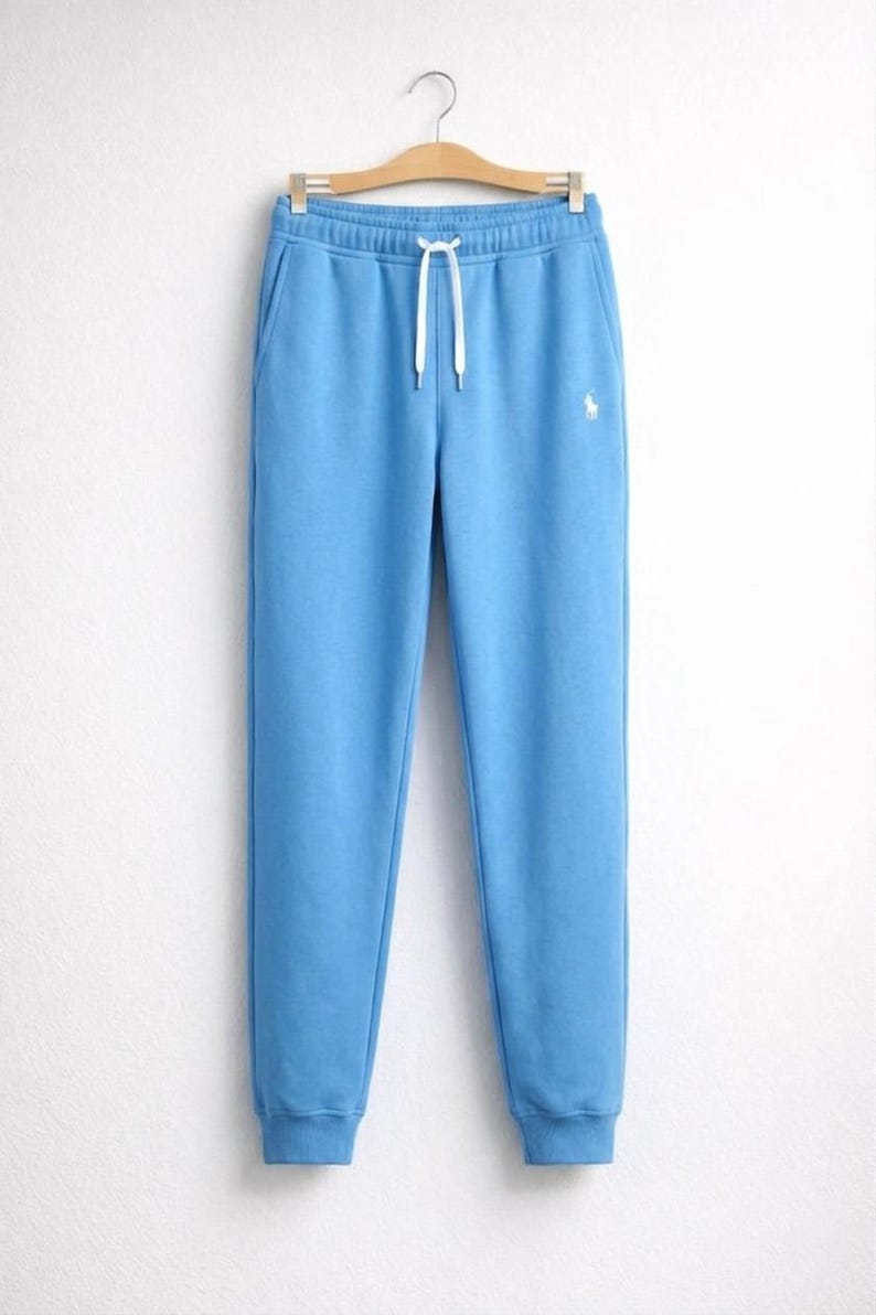 May include: Light blue sweatpants with a drawstring waist and a small white logo on the left leg. The pants have side pockets and are made of a soft material. The sweatpants are hanging on a wooden hanger.