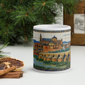 Puede incluir: Una taza de cerámica blanca con una ilustración detallada del Puente Romano y la Mezquita-Catedral de Córdoba, España. El diseño incluye el texto CORDVBA, enmarcado por un borde decorativo azul y dorado en los bordes superior e inferior.