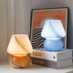 May include: Two mushroom-shaped glass lamps with a marbled pattern. The left lamp emits a warm amber glow, while the right lamp emits a cool blue light. They are positioned on top of two stacked books titled Design Hotels Yearbook 09 and Jed Lind.