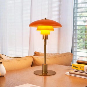 May include: A table lamp featuring a bright orange, multi-tiered shade structure with a glowing yellow inner light source. The lamp has a slender, metallic gold-toned stem and a circular base, standing upright on a wooden surface.