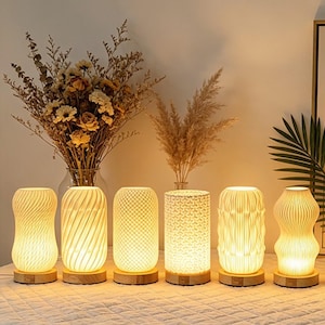 May include: Six cylindrical table lamps with white textured shades and light wood bases. Each lamp features a unique geometric pattern, including vertical waves, diagonal swirls, cross-hatching, and intricate repeating shapes, emitting a warm glow.