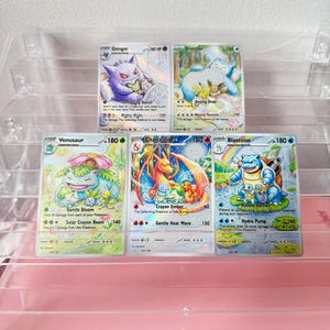 May include: Five Pokémon trading cards displayed in a clear plastic tiered stand. The cards feature Gengar, Snorlax, Venusaur, Charizard, and Blastoise with colorful, detailed illustrations of the characters in various scenes with text and stats.