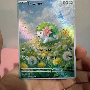 Può includere: Una carta collezionabile Pokémon Shaymin con 80 HP, raffigurante una creatura verde e bianca con fiori rosa in un campo di denti di leone. La carta mostra gli attacchi Flower Dash e Breeze Blessing, con una finitura olografica brillante.