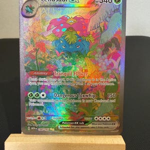 May include: A holographic Venusaur EX Pokémon card. The card shows a colourful illustration of Venusaur in a jungle setting. Text includes "Venusaur EX", "Tranquil Flower", and "Dangerous Toxwhip". The card is displayed on a small wooden easel.