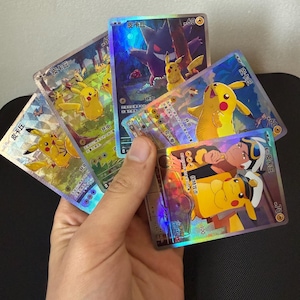 May include: A hand holding a fan of holographic trading cards featuring the character Pikachu. The cards have Japanese text and various illustrations of Pikachu in different poses and settings. The cards have a shiny, reflective surface.