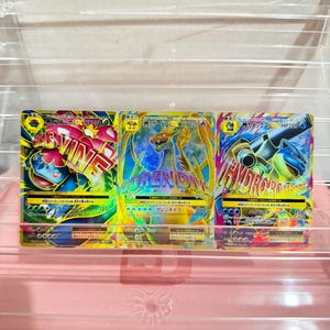 Puede incluir: Tres cartas coleccionables Pokémon japonesas con Mega Venusaur, Mega Charizard y Mega Blastoise. Cada carta muestra ilustraciones vibrantes, texto en japonés, valores de HP de 230 o 220 y nombres de ataques en negrita como Crisis Vine, Guren Dive y Hydro Bomb.