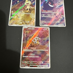 Op de afbeelding: Drie Pokémon-verzamelkaarten met kleurrijke illustraties. Eén kaart toont een Meowth, een andere een Gengar en de derde een Cubone. Elke kaart bevat Japanse tekst en numerieke waarden.