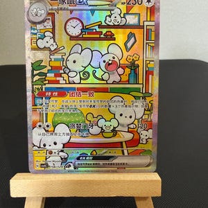 Op de afbeelding: Een holografische ruilkaart met cartoonmuizen in een kleurrijke, gestileerde setting. De kaart, met Japanse tekst, toont muizen met boeken, klokken en andere objecten. De kaart wordt getoond op een kleine houten ezel.