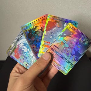 Może przedstawiać: Cztery holograficzne karty kolekcjonerskie Pokémon są trzymane w dłoni. Karty przedstawiają kolorowe ilustracje różnych postaci Pokémon, w tym Solgaleo & Lunala i Reshiram & Zekrom. Każda karta ma etykietę "TAG TEAM" i oznaczenie "GX".