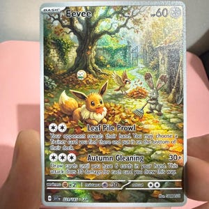 May include: A collectible Eevee trading card featuring a detailed autumn forest scene with Eevee sitting in fallen leaves, a Buneary raking leaves, and a Pidgey nearby. The card displays the text Leaf Pile Prowl and Autumn Gleaning with HP 60.