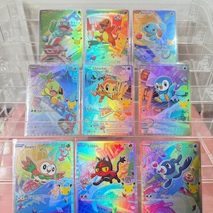 May include: A collection of nine holographic Pokémon trading cards arranged in a clear plastic display stand. Each card features a colorful illustration of a different creature, including Bulbasaur, Charmander, Squirtle, Turtwig, Chimchar, Piplup, Rowlet, Litten, and Popplio, all with a distinct rainbow foil finish and a small Pikachu icon.