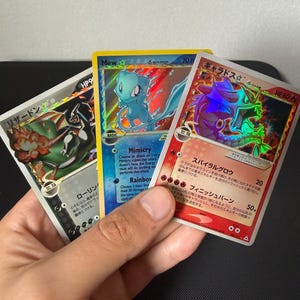 May include: Three Pokémon trading cards are held in a hand. The cards feature a Charizard, Mew, and Gyarados. The cards have holographic foil and Japanese text. The Mew card is blue, the Gyarados card is red, and the Charizard card is black and white.