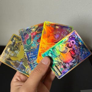 Peut inclure: Quatre cartes Pokémon holographiques sont tenues dans une main. Les cartes présentent divers personnages, dont Pikachu, Dracaufeu et Tortank. Chaque carte a un design et une palette de couleurs différents, avec du texte japonais et des valeurs HP.