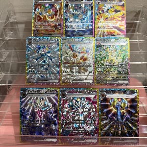 May include: A collection of nine holographic trading cards arranged in a three-by-three grid on a clear plastic display stand. Each card features colourful Japanese text, intricate character illustrations, and shiny, reflective foil patterns.