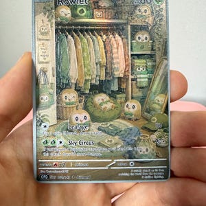 May include: A Rowlet trading card featuring a detailed illustration of the character in a closet filled with clothes, accessories, and multiple Rowlet figures. The card displays the name Rowlet, 60 HP, attacks Leafage and Sky Circus, and text including small but mighty.