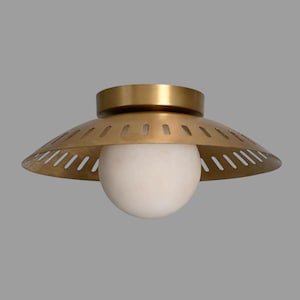 May include: A flush mount ceiling light fixture featuring a wide, circular brass shade with a series of evenly spaced vertical cutouts along the rim. A large, spherical white glass globe sits centered beneath the metallic brass canopy.