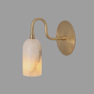May include: A wall sconce featuring a circular brass backplate and a curved brass arm that supports a cylindrical, translucent white stone shade with natural veining. The light fixture emits a soft glow through the stone material.
