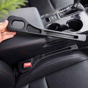 Puede incluir: Guía de cinturón de seguridad y accesorio de reposabrazos de plástico negro para automóvil. La guía tiene un diseño curvo con un asa y una ranura. El reposabrazos es negro con costuras rojas. El interior del coche es negro.