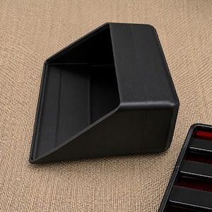 May include: A matte black plastic storage bin with an angled front opening and a rectangular base. Beside it lies a separate black tray component featuring horizontal slats and a dark red interior lining, both designed for organized storage.