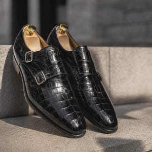 Puede incluir: Un par de zapatos de doble hebilla en color negro con una textura de cuero brillante con relieve de cocodrilo, hebillas met&aacute;licas plateadas y hormas de madera clara insertadas en la abertura de cada zapato.