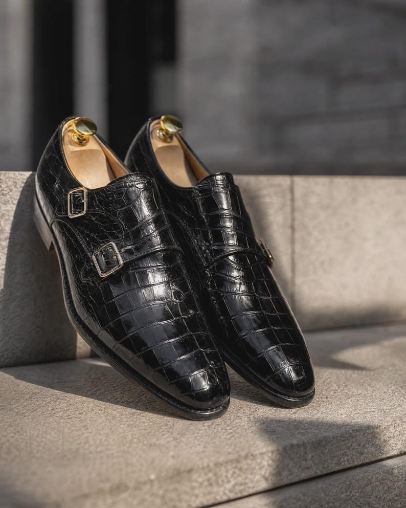 Puede incluir: Un par de zapatos de doble hebilla en color negro con una textura de cuero brillante con relieve de cocodrilo, hebillas met&aacute;licas plateadas y hormas de madera clara insertadas en la abertura de cada zapato.