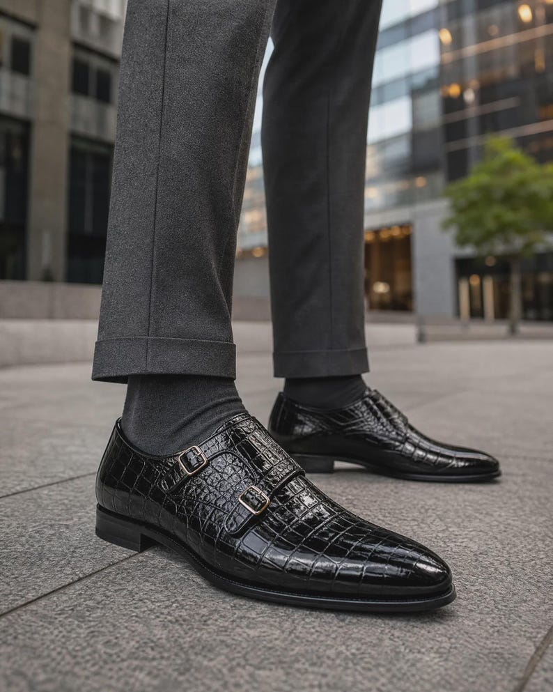 Puede incluir: Un par de zapatos de doble hebilla en color negro con una textura brillante de relieve de cocodrilo, con dos hebillas met&aacute;licas en el lateral, usados con pantalones gris oscuro y calcetines a juego, con punta afilada y tac&oacute;n bajo.