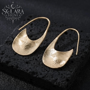 May include: A pair of gold-toned earrings featuring a curved, open-loop design with a textured, hammered surface. The earrings have a smooth, polished finish on the edges and a distinct, irregular pattern across the front, creating a reflective look.