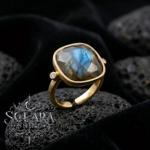 May include: A gold ring featuring a large, square-cut labradorite gemstone with a blue iridescent flash. The setting includes two small, round, clear accent stones on either side of the central gem. The band is smooth and polished gold.