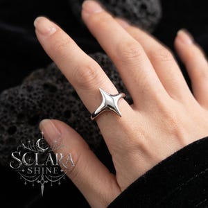 May include: A silver ring with a starburst design. The ring is worn on a finger, showcasing its polished, reflective surface. The ring's design features four pointed extensions radiating from a central point. The ring is a piece of jewelry.